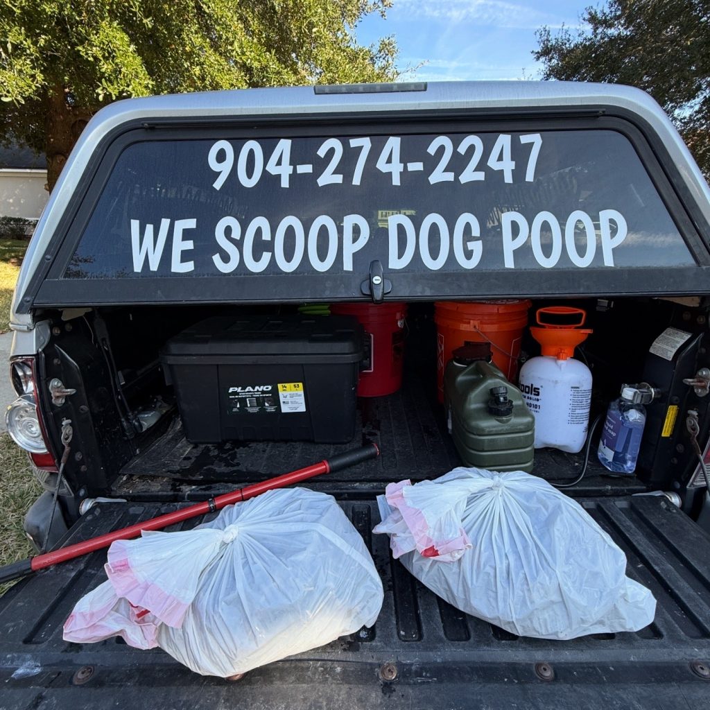 553C7498-2111-48D4-ADC9-FA6E8D9D1098 Bagged pet waste on the back of a tailgate of a truck. The truck cap window says "we scoop dog poop" and the phone number 9042742247 is listed.